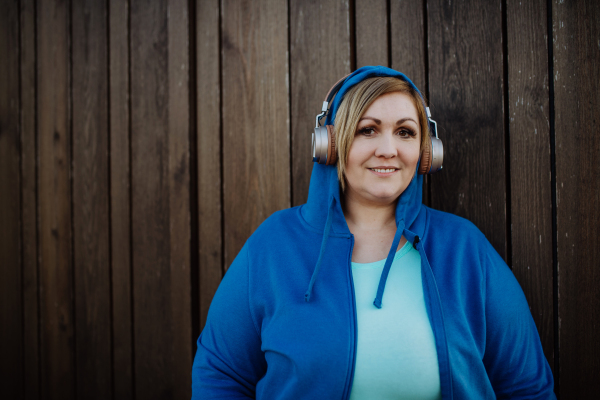 A happy overweight woman in sports clothes with headphones looking at camera against wooden wall