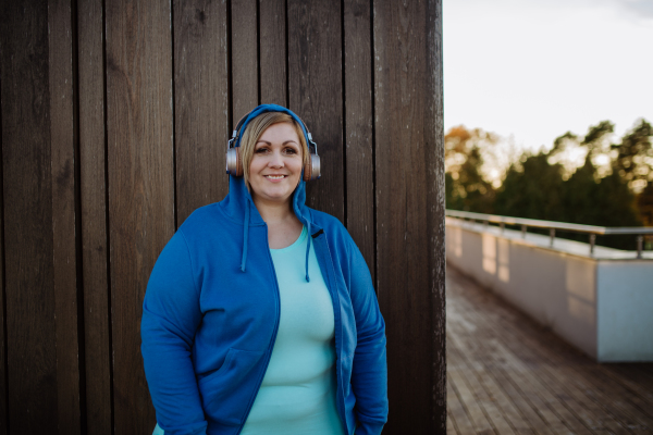 A happy overweight woman in sports clothes with headphones looking at camera against wooden wall