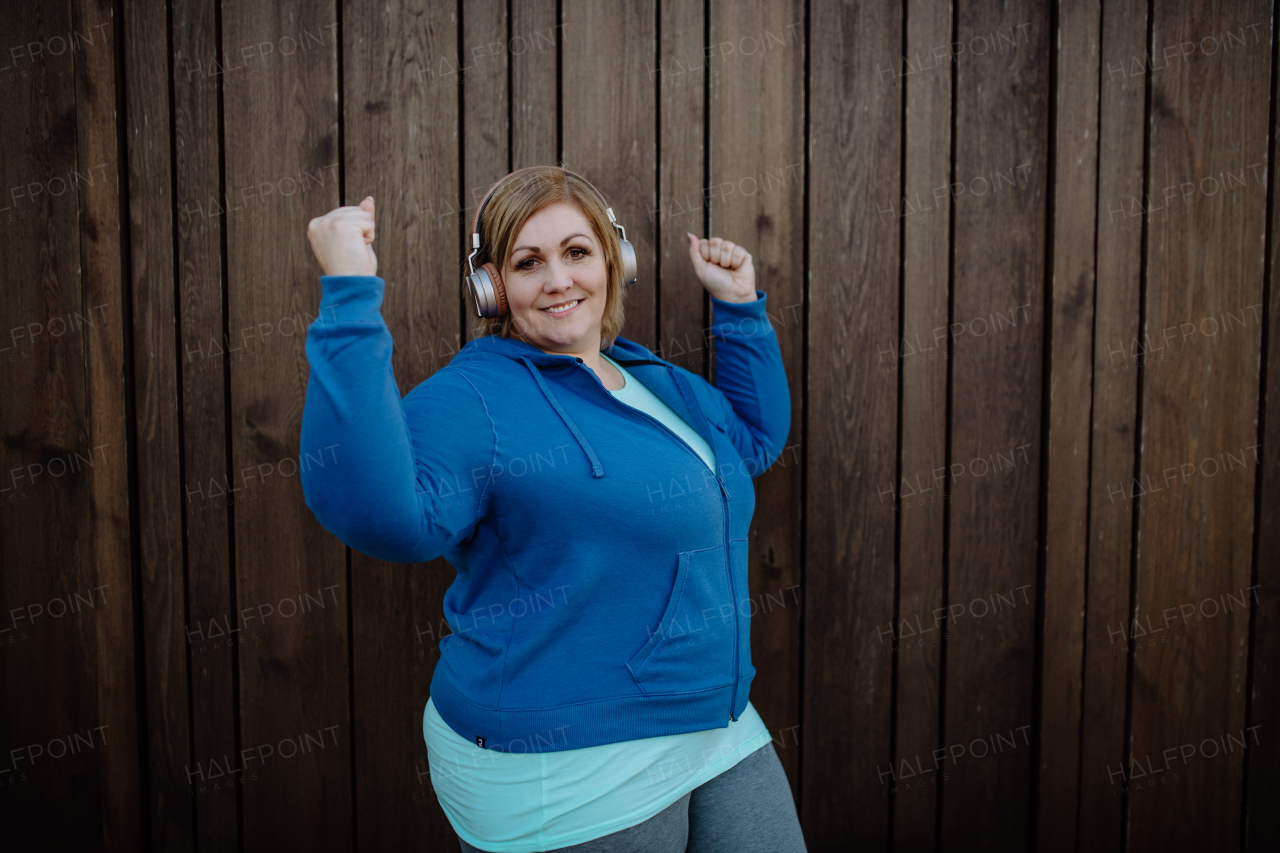 A happy overweight woman in sports clothes with headphones looking at camera against wooden wall