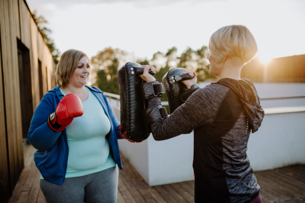 An overweight woman training boxing with personal trainer outdoors on terrace.