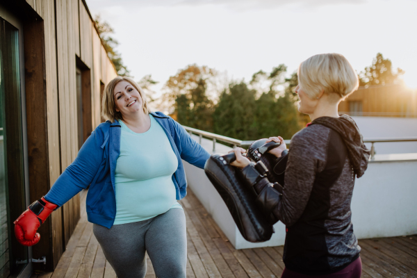 An overweight woman training boxing with personal trainer outdoors on terrace.