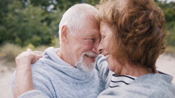 A front view of happy senior couple in love on walk outdoors on beach at morning, hugging.