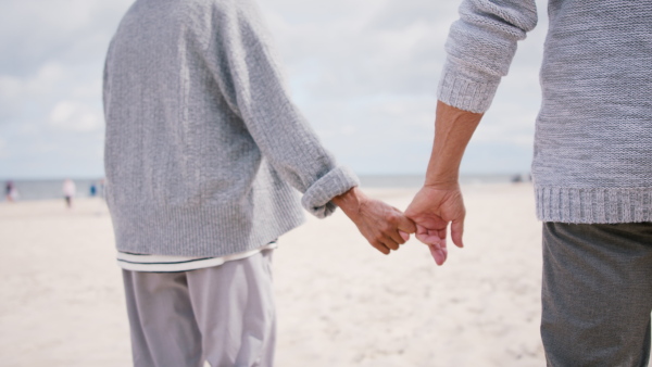 A rear view of happy senior couple in love on walk outdoors on beach at morning, close up of holding hands.