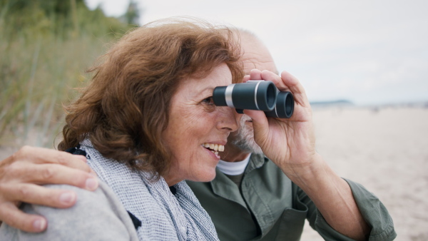 A happy senior couple outdoors on beach, looking at view through binoculars.