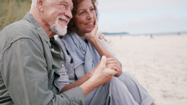 A happy senior couple outdoors on beach, holding hands, sitting and hugging.