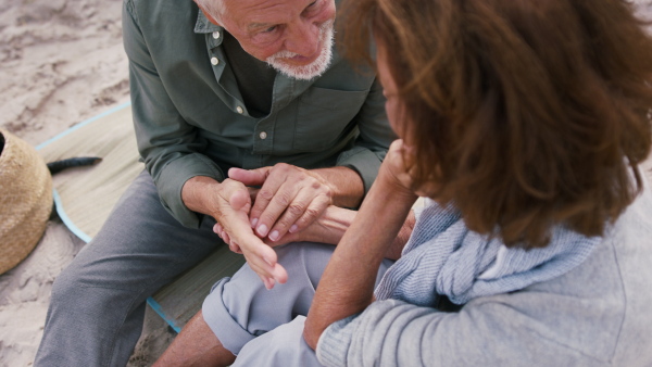 A happy senior couple outdoors on beach, holding hands and sitting.