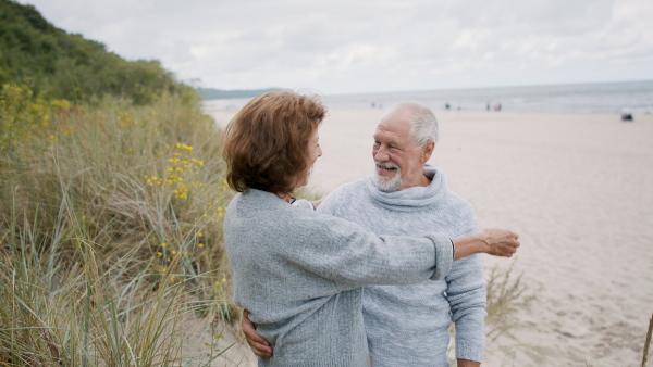 A front view of happy senior couple in love on walk outdoors on beach at morning, hugging.