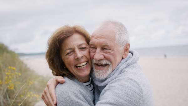 A front view of happy senior couple in love on walk outdoors on beach at morning, hugging.