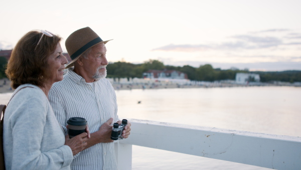 A happy senior couple hugging outdoors on pier by sea, looking at view through binoculars.