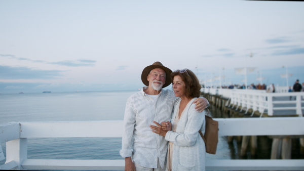 A happy senior couple outdoors on pier by sea, posing for photo.