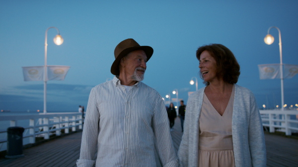 A happy senior couple in love on walk outdoors on pier by sea at dusk, holding hands.