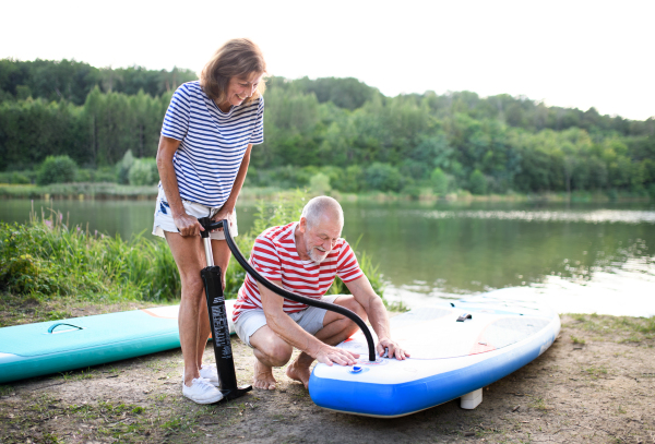 Active senior couple getting ready for paddleboarding by lake in summer.