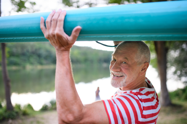 Portrait of senior man carrying paddleboard by lake in summer, looking at camera.