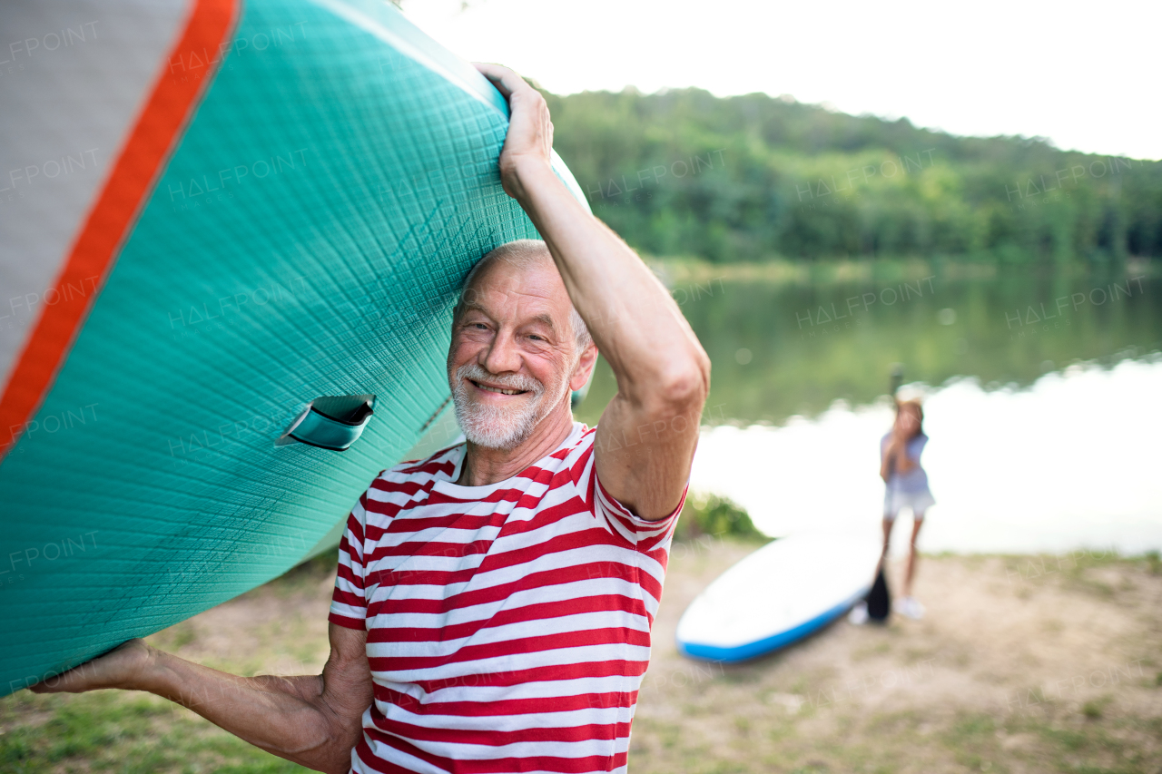 Portrait of senior man carrying paddleboard by lake in summer, looking at camera.