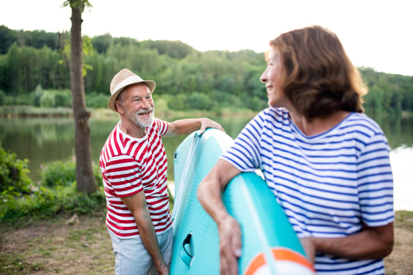 Portrait of senior man carrying paddleboard by lake in summer, talking.