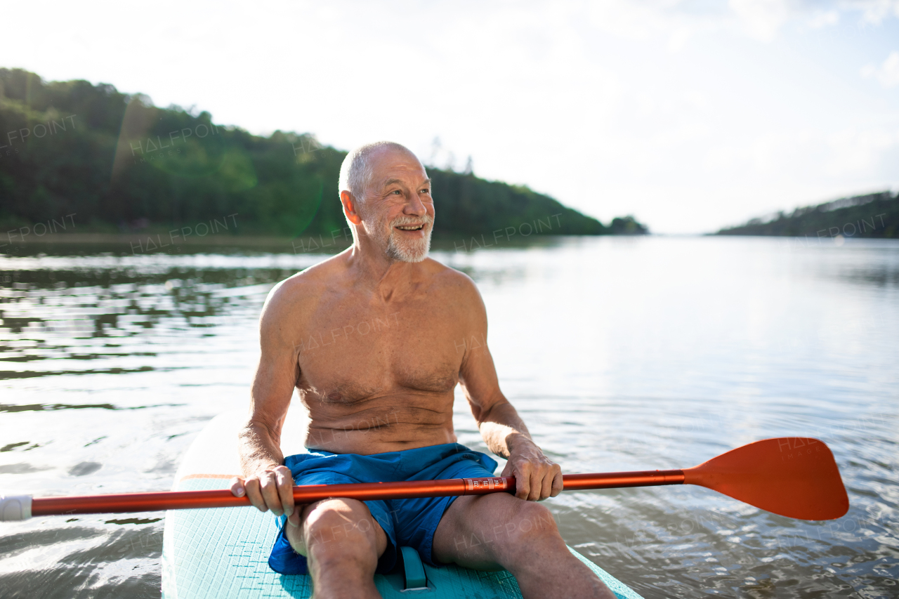 Front view of senior man sitting on paddleboard on lake in summer. Copy space.