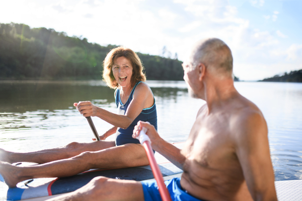 Active senior couple paddleboarding on lake in summer.