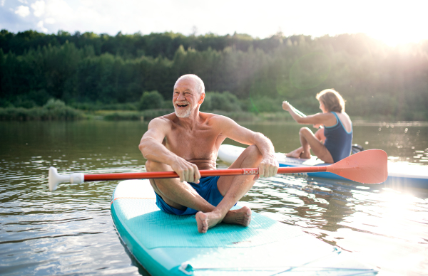 Active senior couple paddleboarding on lake in summer.