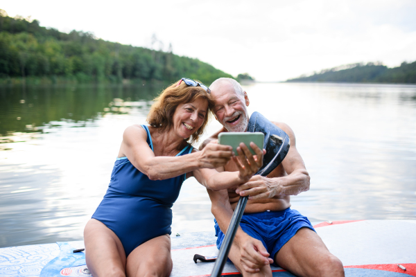 Cheerful senior couple sitting on paddleboard on lake in summer, taking selfie with smartphone.