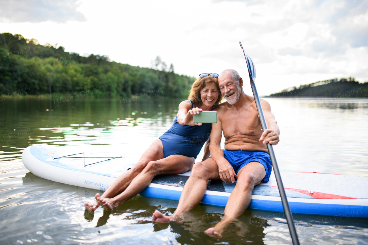 Cheerful senior couple sitting on paddleboard on lake in summer, taking selfie with smartphone.