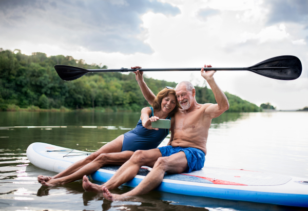 Cheerful senior couple sitting on paddleboard on lake in summer, taking selfie with smartphone.