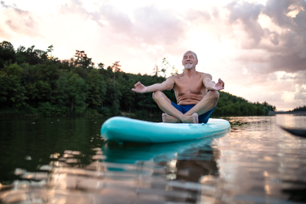 Senior man on paddleboard on lake in summer, doing yoga meditation exercise.