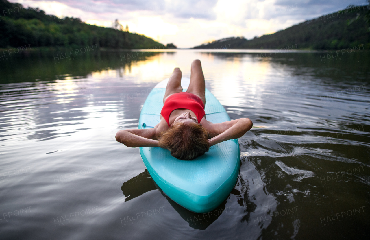 Rear view of contented senior woman lying on paddleboard on lake in summer, resting.