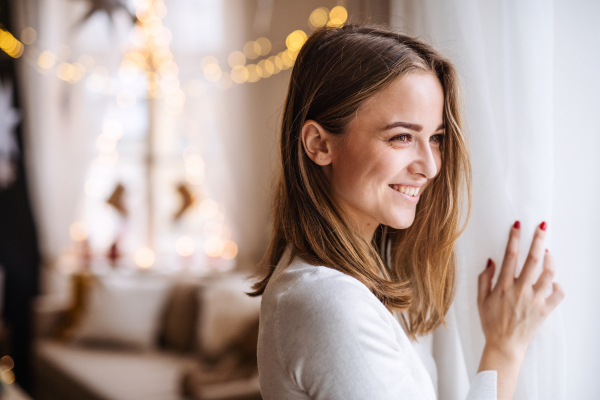 Portrait of happy young woman indoors at home at Christmas, looking out of window.