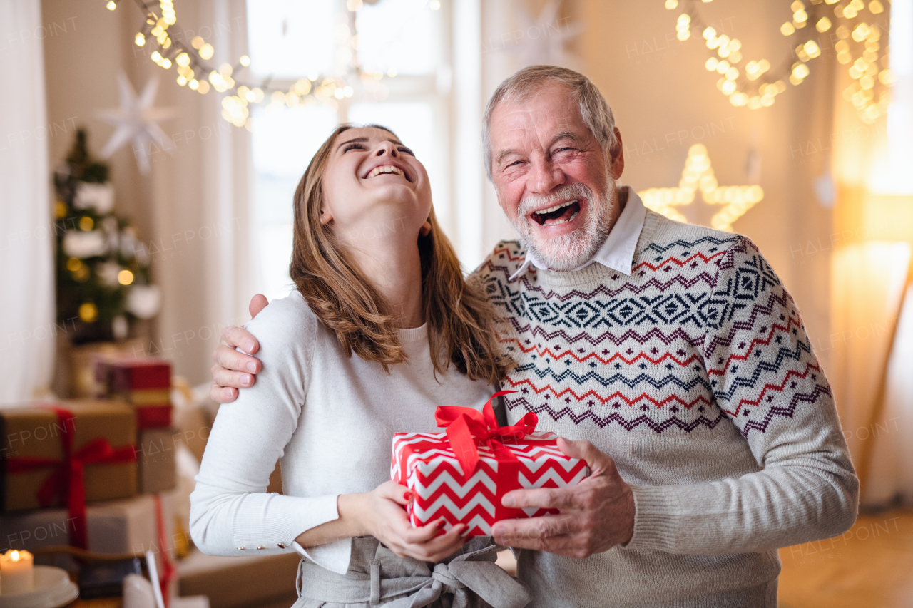 Laughing senior man with young woman indoors at home at Christmas, holding present.