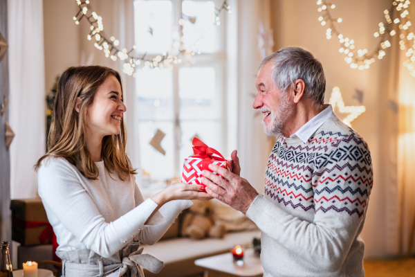 Portrait of senior man giving present to happy granddaughter indoors at home at Christmas.