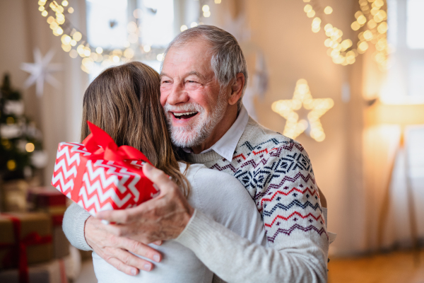 Portrait of young woman giving present to happy grandfather indoors at home at Christmas.