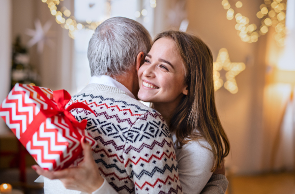 Portrait of young woman giving present to happy grandfather indoors at home at Christmas.