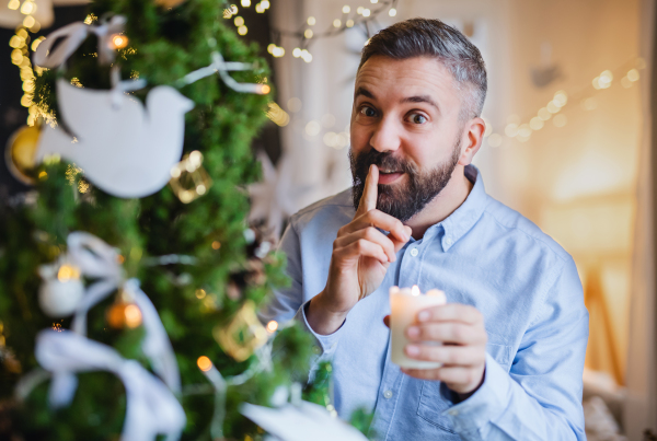 Mature man with candle indoors at home at Christmas, making silence gesture.