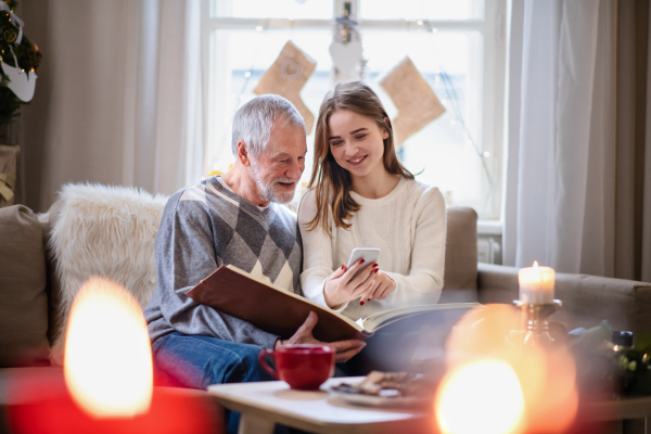 Portrait of young woman with grandfather indoors at home at Christmas, using smartphone.