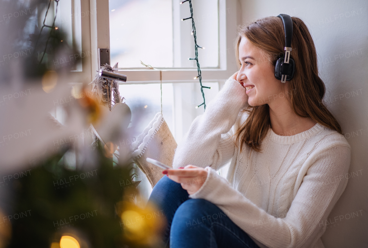 Portrait of young woman sitting indoors at home at Christmas, using smartphone and headphones.