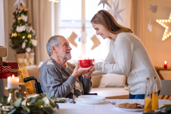 Young woman giving cup of tea to senior grandfather in wheelchair indoors at home at Christmas.