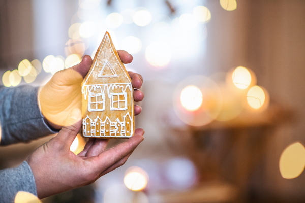 Hands of man holding gingerbread biscuit house indoors at Christmas, close-up. Copy space.