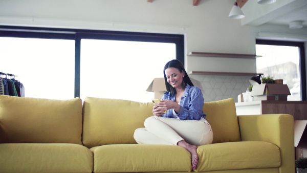 A young woman moving in new home, sitting on sofa and using smartphone. A moving in new home concept.