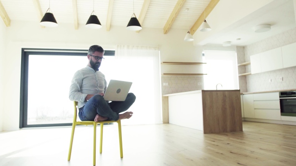 A mature man sitting on chair in unfurnished new house, using laptop. A new home concept.