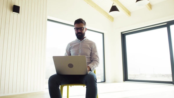 A mature man sitting on chair in unfurnished new house, using laptop. A new home concept.