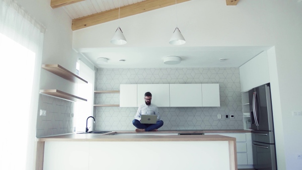 A mature man with laptop sitting on kitchen counter in new house. Moving in new home concept.