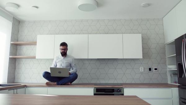 A mature man with laptop sitting on kitchen counter in new house. Moving in new home concept.