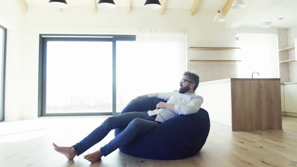 Cheerful mature man sitting on bean bag in unfurnished house, moving in new home concept.