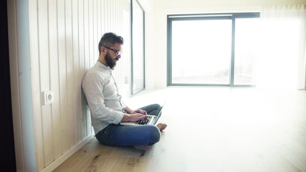 A mature man sitting on the floor in unfurnished new house, using laptop. A new home concept. Slow motion.