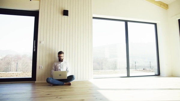 A mature man sitting on the floor in unfurnished new house, using laptop. A new home concept. Slow motion.