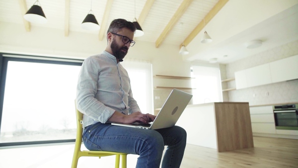 A mature man sitting on chair in unfurnished new house, using laptop. A new home concept. Slow motion.