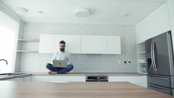 A mature man with laptop sitting on kitchen counter in new house. Moving in new home concept. Slow motion.