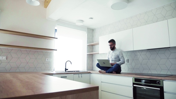 A mature man with laptop sitting on kitchen counter in new house. Moving in new home concept. Slow motion.