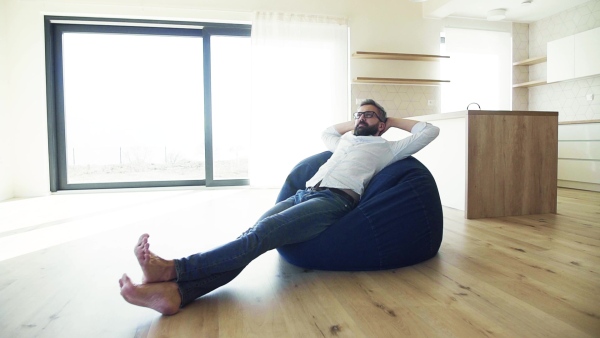 Cheerful mature man jsitting on bean bag in unfurnished house, moving in new home concept. Slow motion.