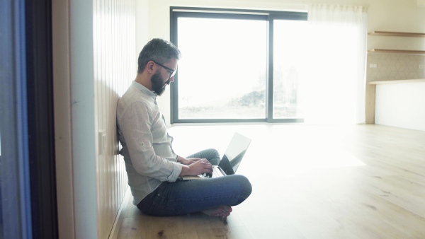 A mature man sitting on the floor in unfurnished new house, using laptop. A new home concept. Copy space.